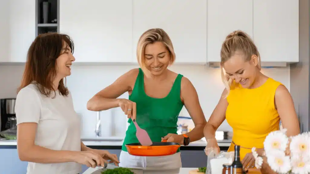 Three women cooking together in a modern kitchen with white cabinets and stainless steel appliances