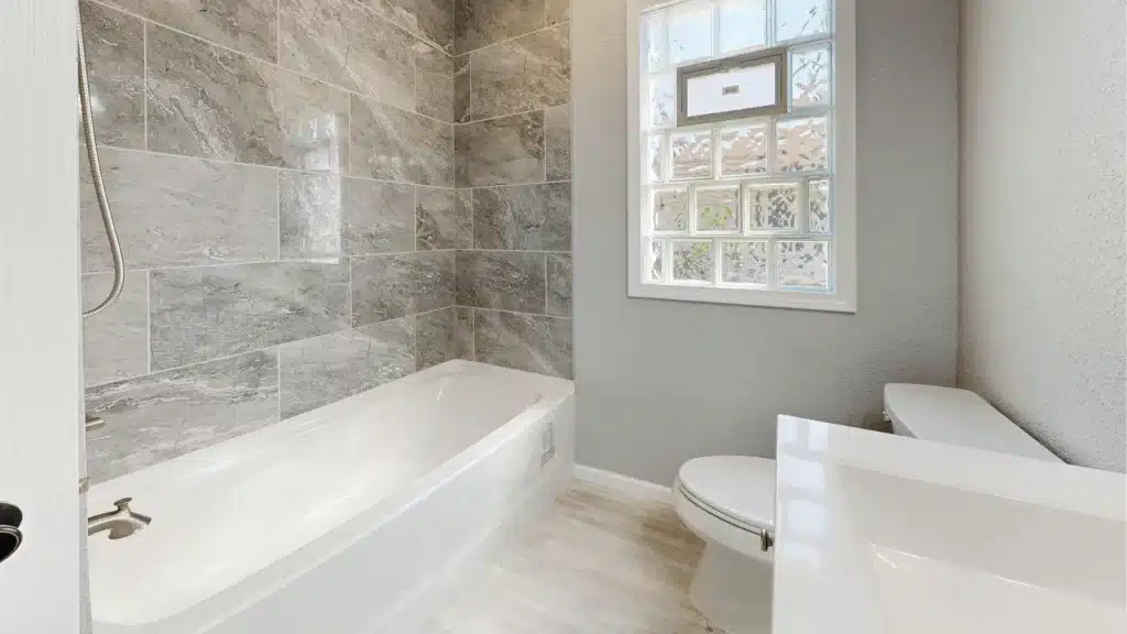 Modern bathroom with travertine tile shower, white bathtub, toilet, and light wood flooring by Pottstown Construction