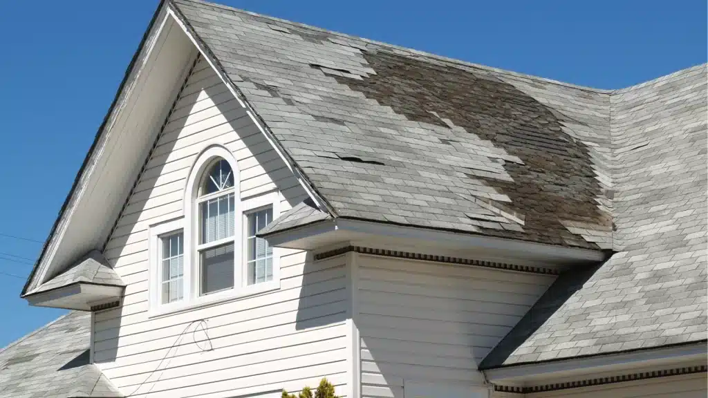 Damaged roof with missing shingles on white sided house requiring roof repair and replacement