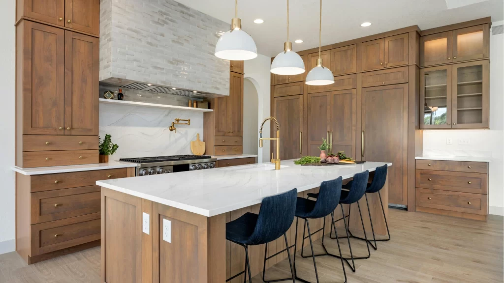 High-end kitchen with warm walnut wood cabinetry, white quartz island, marble stone backsplash, brass fixtures, and navy blue bar stools
