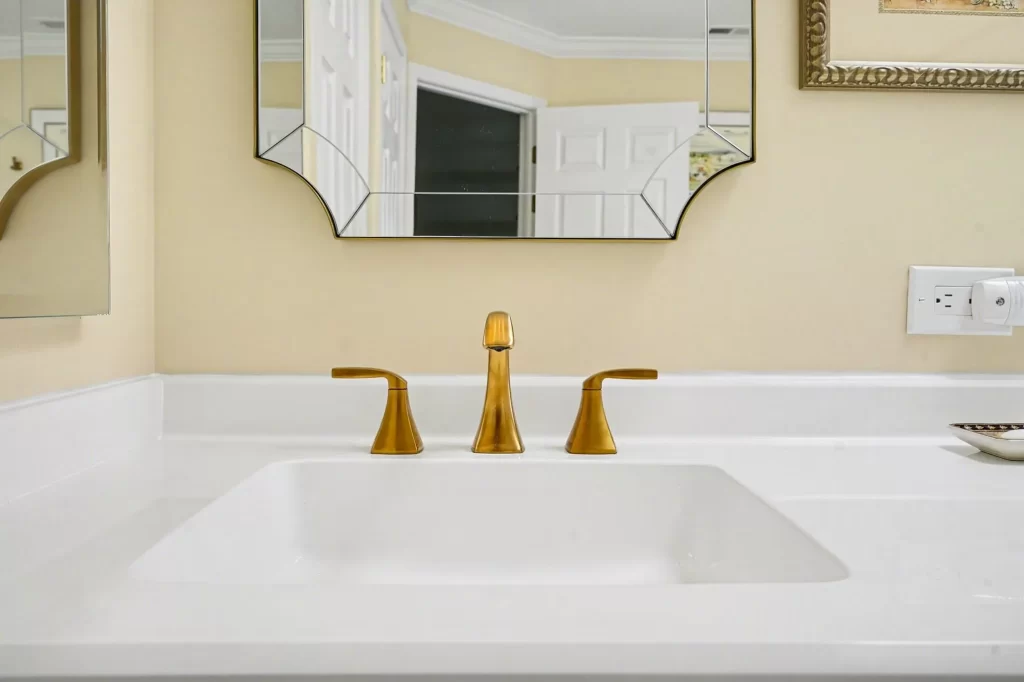 Bathroom sink detail with white quartz countertop, three-piece gold faucet, and octagonal gold-framed mirror on pale yellow wall