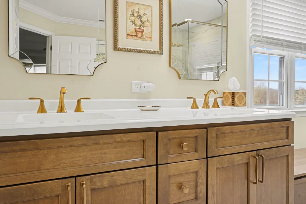 Bathroom vanity detail with honey-wood cabinetry, white quartz countertop, dual gold faucets, and ornate framed mirrors on pale yellow wall.