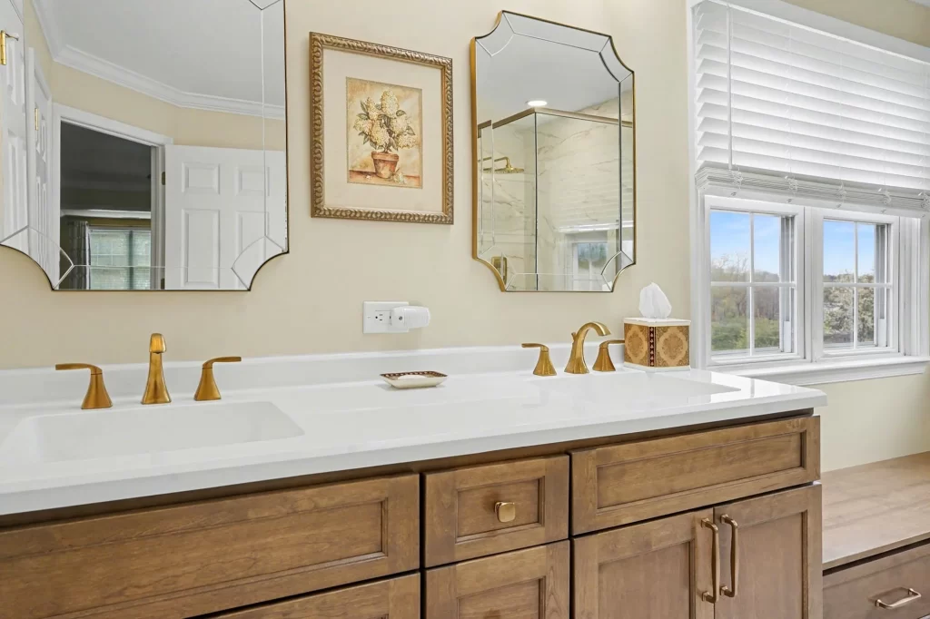 Bathroom vanity with honey-wood cabinetry, white quartz countertop, dual gold faucets, ornate framed mirrors, and windows with scenic views.