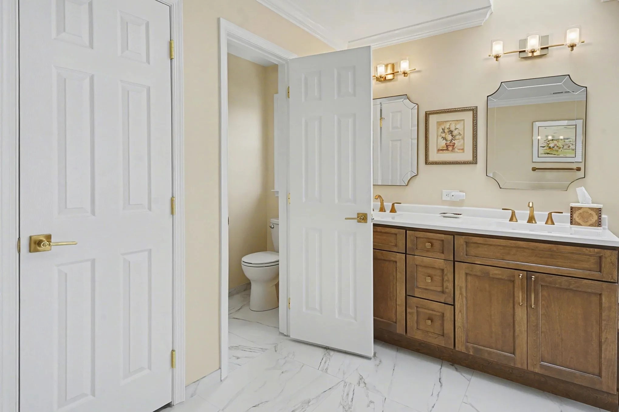 Spacious master bathroom with double vanity, honey-wood cabinetry, white quartz countertop, decorative mirrors, and marble flooring with pale yellow walls.