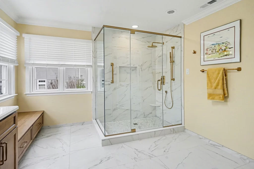 Luxury walk-in shower with marble walls and floors, frameless glass doors, gold fixtures, and large window with pale yellow bathroom walls.
