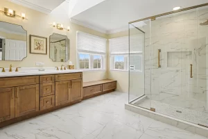 Master bathroom with double vanity, walk-in shower, marble floors and walls, honey-wood cabinetry, and large windows overlooking scenic views.