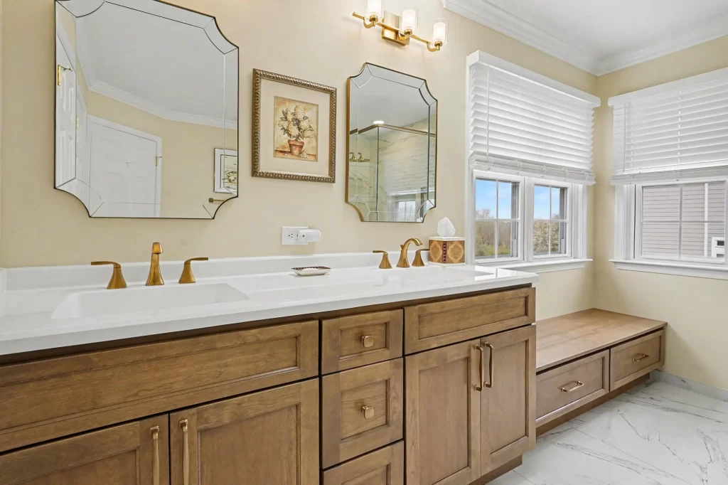 Master bathroom with long double vanity, ornate gold-framed mirrors, honey-wood cabinetry, and built-in window bench with pale yellow walls.