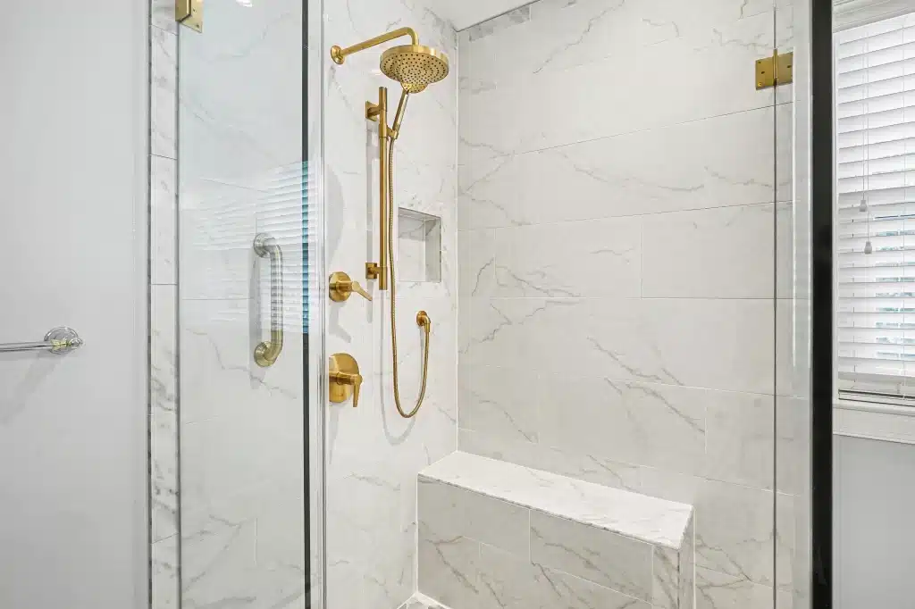 Shower detail with marble-look wall tile, brass fixtures, and built-in bench