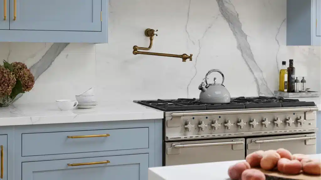 Blue-gray kitchen with a brass pot filler on a white marble slab backsplash above a professional-style gas range with gold cabinet hardware