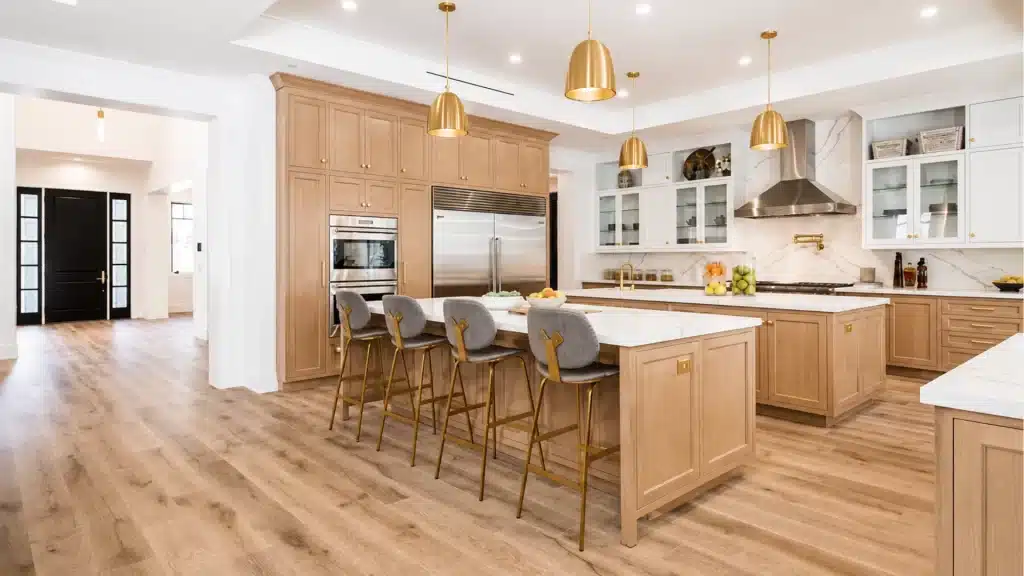 Double-island kitchen with light wood cabinets, gold pendant lights, and white marble backsplash