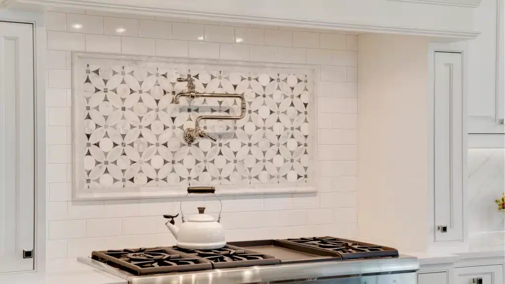 Chrome pot filler mounted on a decorative marble mosaic tile backsplash above a stainless steel gas range with a white kettle on the cooktop
