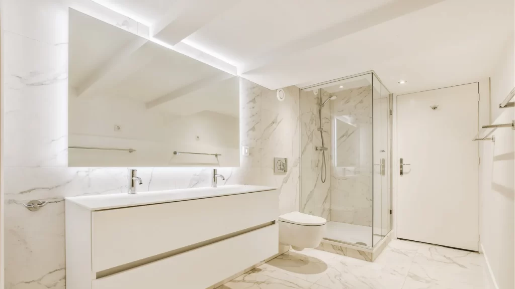 All-white bathroom with a large backlit LED mirror over a floating double vanity, glass shower, and marble tile flooring
