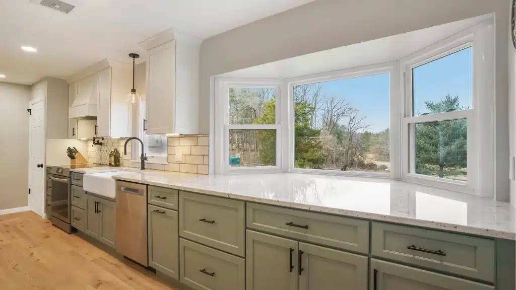 Modern kitchen remodel with green cabinets, white quartz countertops, farmhouse sink, and large windows overlooking trees