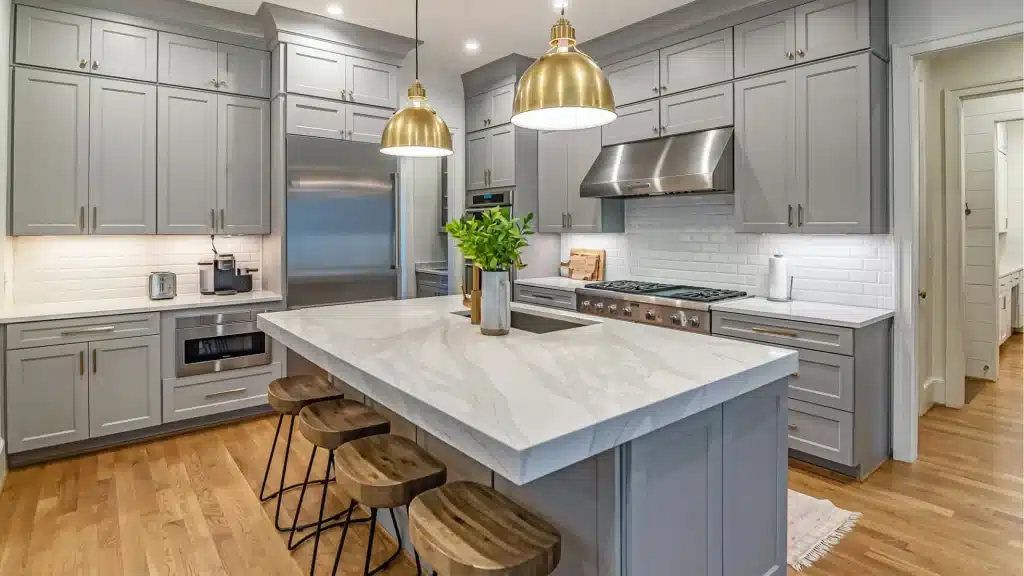 Modern kitchen renovation with gray shaker cabinets, marble island countertops, brass pendant lights, and stainless steel appliances