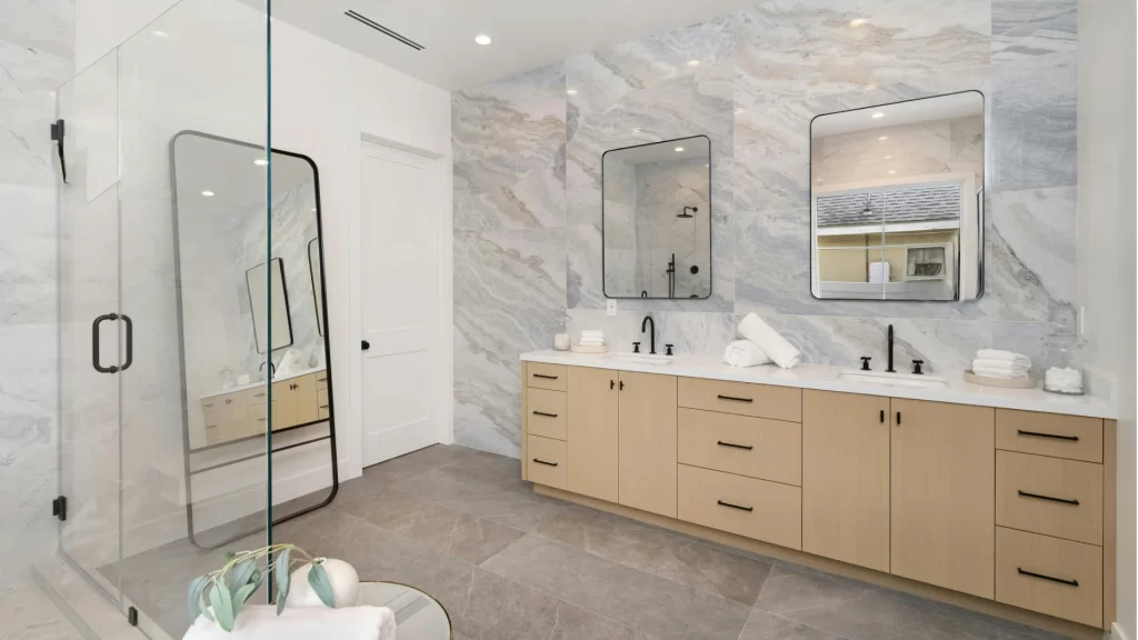 Modern luxury bathroom with marble tile walls, wood vanity cabinets, frameless glass shower, and dual sinks