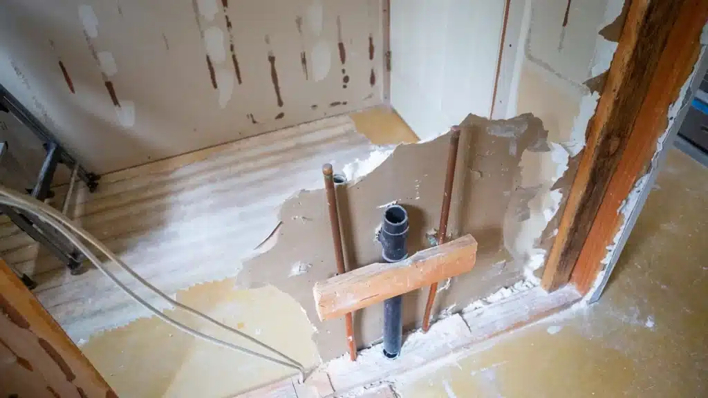 Older home bathroom gutted to the studs, exposing copper supply lines, a black drain pipe, and worn subfloor during remodel