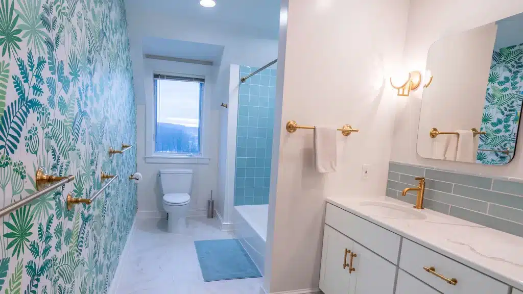 Bathroom with tropical leaf wallpaper, blue subway tile tub surround, gold towel bars, and white quartz vanity